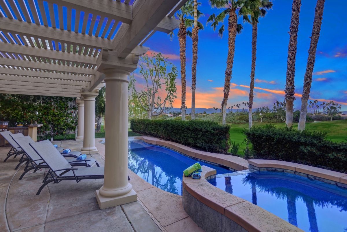 8cfef338-aa28-4b2b-885a-8682672fb81b | Oranj Palm Vacation Homes Backyard pool area at sunset with lounge chairs under a pergola and tall palm trees nearby.