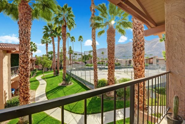 Balcony view of a sunny resort courtyard with palm trees, green lawn, and a fenced tennis court, mountains in the distance.