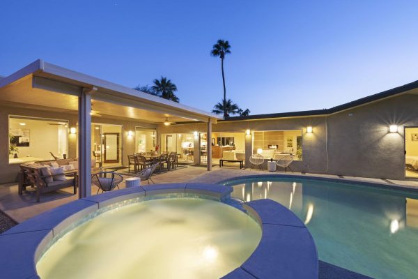 Backyard of a modern house at dusk with a lit patio, seating, outdoor dining, a circular hot tub and a pool.