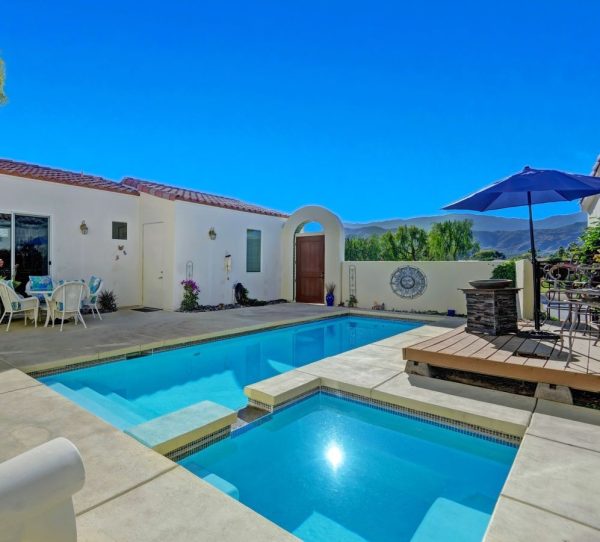 Sunny backyard with a turquoise rectangular pool and raised spa, surrounded by a concrete patio, white stucco house, and outdoor seating under a large blue umbrella with mountain views in the distance.